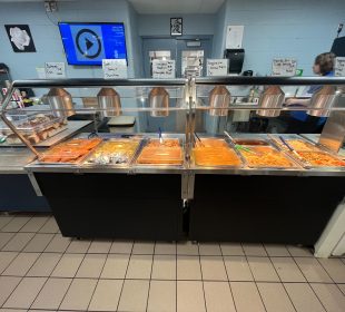 A buffet of 11 food choices with a bucket of buns off to the side. There is a server behind the buffet.