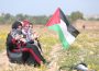 Four people sit in a field of dandelions wearing traditional Palestinian clothing. The third person of the four holds a Palestine flag out to billow in the wind.
