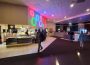 An image of the concession area of Moonlight Movies. There are some people wandering and one person working the register behind the counter. There are some chairs and tables and some arcade games at the back of the image.