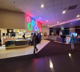 An image of the concession area of Moonlight Movies. There are some people wandering and one person working the register behind the counter. There are some chairs and tables and some arcade games at the back of the image.