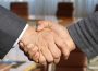 A handshake between two masculine-appearing hands, one showing a suit coat cuff and the other a casual sweater cuff.