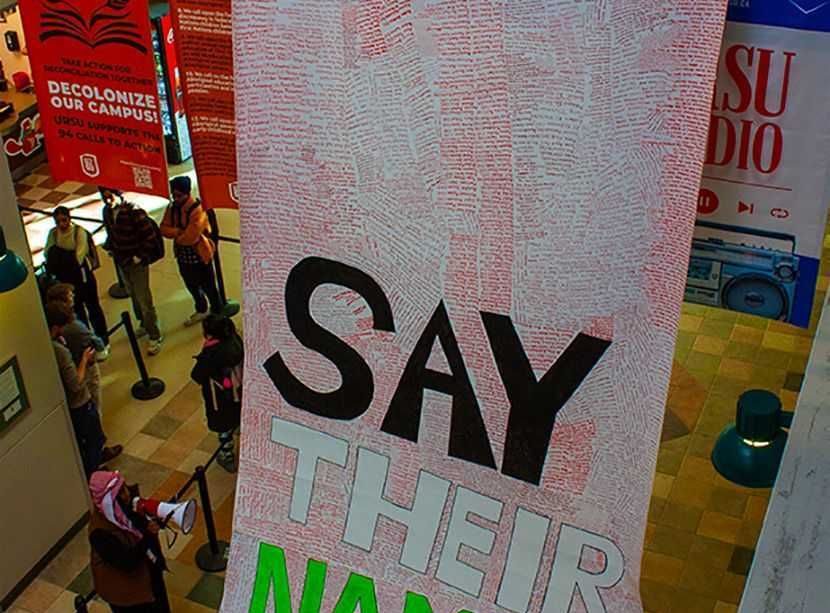A photograph of a massive banner filled with handwritten names of roughly 5,000 of the murdered in Palestine drapes from the second story and across the lower-level floor titled “Say Their Names.”