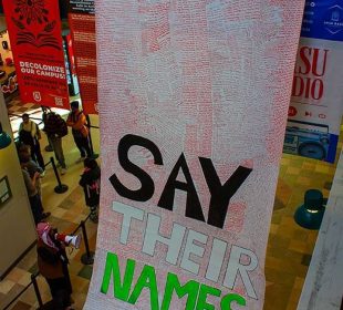 A photograph of a massive banner filled with handwritten names of roughly 5,000 of the murdered in Palestine drapes from the second story and across the lower-level floor titled “Say Their Names.”
