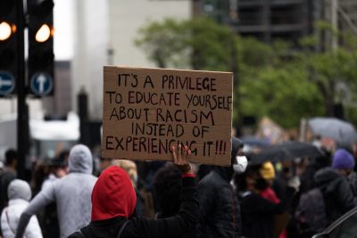 Black person in red hood holds sign that reads “It’s a privilege to learn about racism instead of experiencing it!!!”