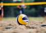 Close up of a blue, yellow, and white volleyball placed in the sand with blurry players and net in the background