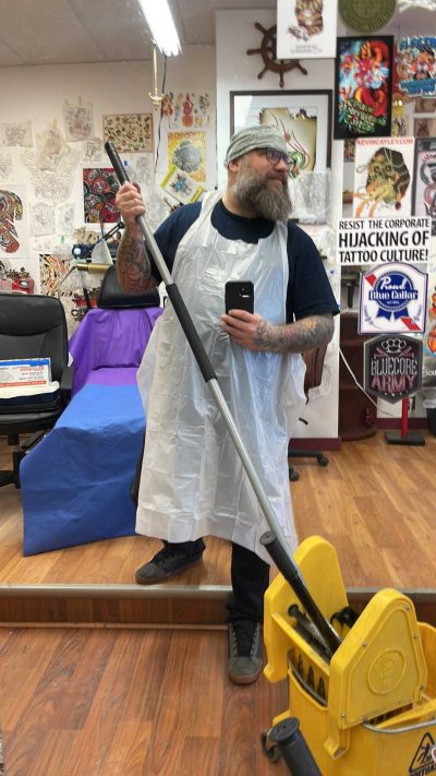 This is a selfie of the owner in his shop. He is wearing a white apron, his arms are covered in tattoos, he has a medium beard and glasses. He also is holding a mop in the picture, and the walls in the background are covered in collected art pieces.