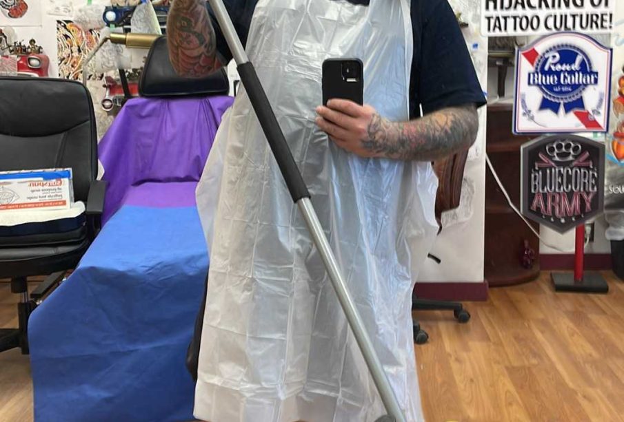 This is a selfie of the owner in his shop. He is wearing a white apron, his arms are covered in tattoos, he has a medium beard and glasses. He also is holding a mop in the picture, and the walls in the background are covered in collected art pieces.