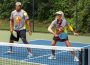 Two people stand on the same side of a pickleball court, with the one on the right having just returned the ball to the other side.