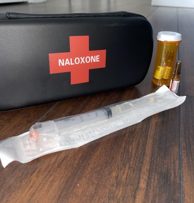 A black naloxone kit sits on a wooden table, with a still-packaged needle and three vials of the liquid injection sit beside it.
