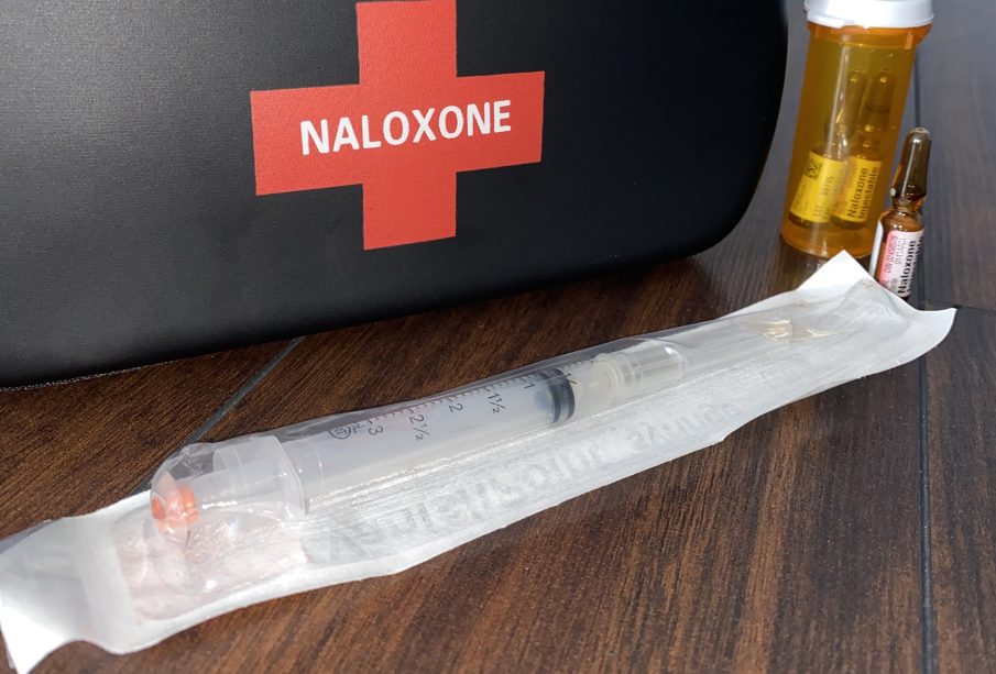 A black naloxone kit sits on a wooden table, with a still-packaged needle and three vials of the liquid injection sit beside it.