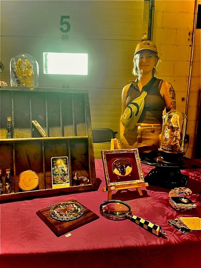 Carrie Sweeney standing to the right behind a table of her creations. There are butterflies encased in intricate wooden frames; a fox skull surrounded by orange, brown, and beige flowers typically found in a meadow encased in a glass dome; another glass dome with a butterfly on a mushroom; a lichen-covered fox skull; The Moon tarot card; and glass vials of butterfly wings with moss. There is also a magnifying glass, a small rabbit figurine, and a circle of wood also on display.