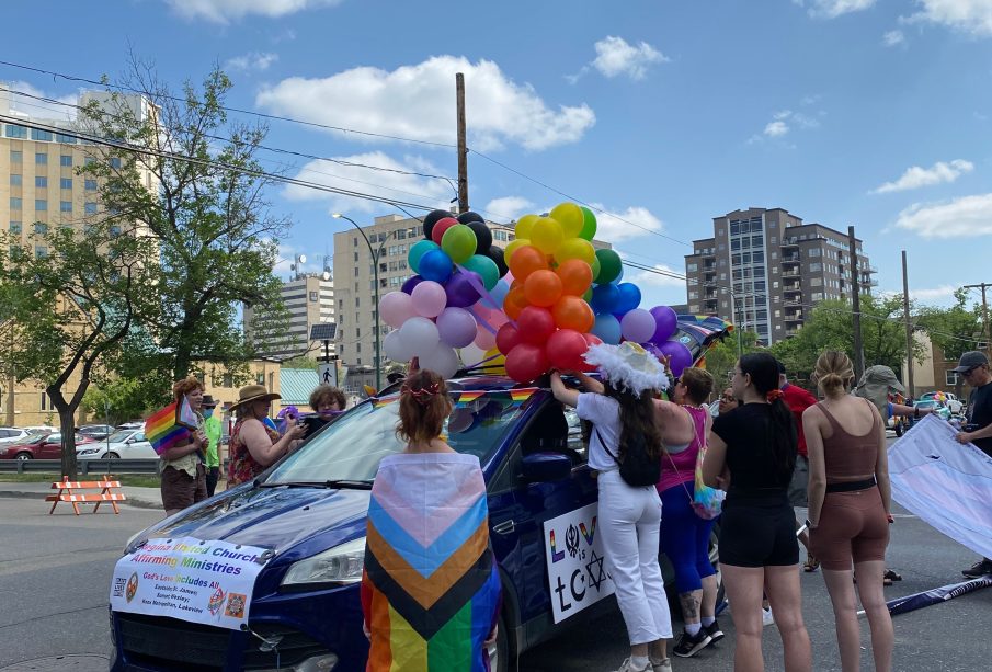 A person is wrapped in a Progressive Pride Flag (the one with a multicolor chevron-style intersection on the left of the rainbow flag) and standing near a group of people decorating a vehicle for the Pride Parade.