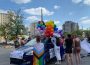 A person is wrapped in a Progressive Pride Flag (the one with a multicolor chevron-style intersection on the left of the rainbow flag) and standing near a group of people decorating a vehicle for the Pride Parade.