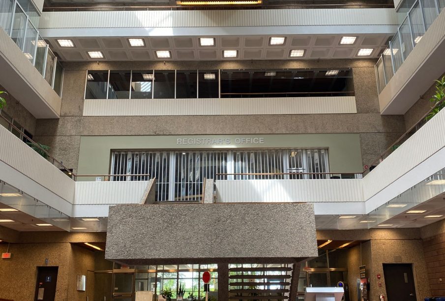 A well-lit, midday photo taken in the ‘pit’ of the Administration Humanities Building facing the main north doors, showing the full height of the building from carpeted floor to the president’s office at the top.