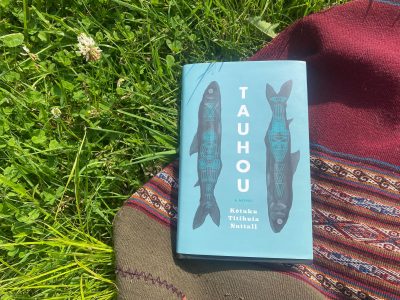A blue book lays on top of a red woven blanket on a patch of grass with some flowers. The book’s cover is its title (Tauhou) written down the middle of the cover in bold black lettering, and the author’s name (Kōtuku Titihuia Nuttall) is written below the title in three lines, and there are two black fish with blue overlaid designs along the sides of the title.