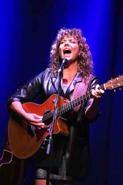 A woman with curly blonde hair is on a stage with a blue backdrop and a white light shining down on her. She is playing a wooden guitar and singing, her head thrown back with the force of her voice.