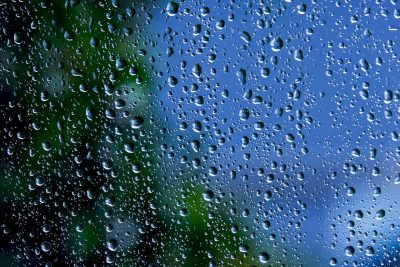 Drops of rain cover a glass pane through which a blue sky and green foliage can be discerned.