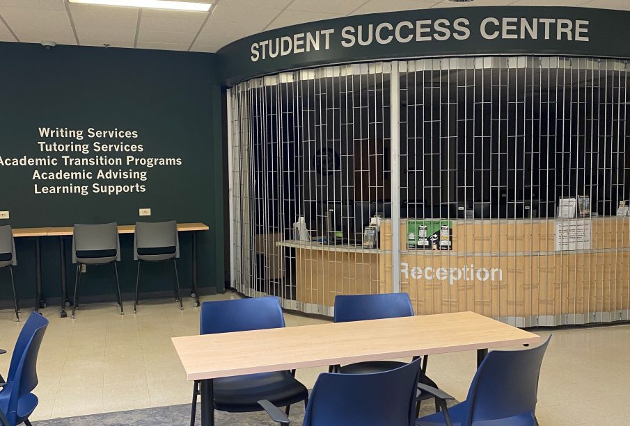 A banner of light-coloured text on a dark background titled Student Success Centre is above a reception area in an open room surrounded by study tables and blue chairs.