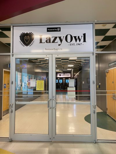 A photo of the double doors to the Lazy Owl at the University of Regina. The doors are made of glass and stainless steel. Above the doors there is a white banner containing a line art image of an owl in a shield and “LazyOwl” in bold, black lettering. Beneath the lettering is “EST. 1967” and the Lazy Owl’s website link: lazyowlyqr.ca.