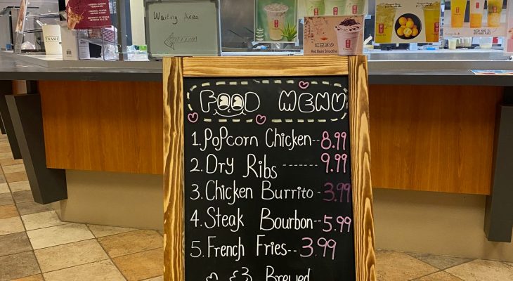 A photo of the entrance to Gong-Cha and its food menu. The menu is a blackboard written on with various colours of chalk, titled “Food Menu” with a smile drawn under the two “o”s in “food”. It contains items such as popcorn chicken for $8.99, dry ribs for $9.99, a chicken burrito $3.99, steak bourbon for $5.99, french fries for $3.99, and a medium brewed coffee for $1.95 or a large for $2.25. There is a drawing of a mug of coffee releasing steam in the shape of a heart and a cloud with “Food available all day” written below it and beside that it says “+ tax,”underlined twice.