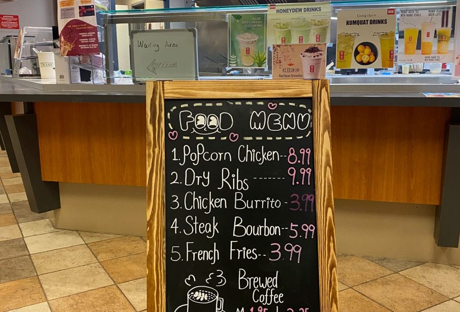 A photo of the entrance to Gong-Cha and its food menu. The menu is a blackboard written on with various colours of chalk, titled “Food Menu” with a smile drawn under the two “o”s in “food”. It contains items such as popcorn chicken for $8.99, dry ribs for $9.99, a chicken burrito $3.99, steak bourbon for $5.99, french fries for $3.99, and a medium brewed coffee for $1.95 or a large for $2.25. There is a drawing of a mug of coffee releasing steam in the shape of a heart and a cloud with “Food available all day” written below it and beside that it says “+ tax,”underlined twice.