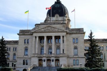 The Saskatchewan Provincial Legislative Building fills the frame, a stone institutional building with a large dome at the centre of the roof and three flags flying from the roof against a white and light blue sky.
