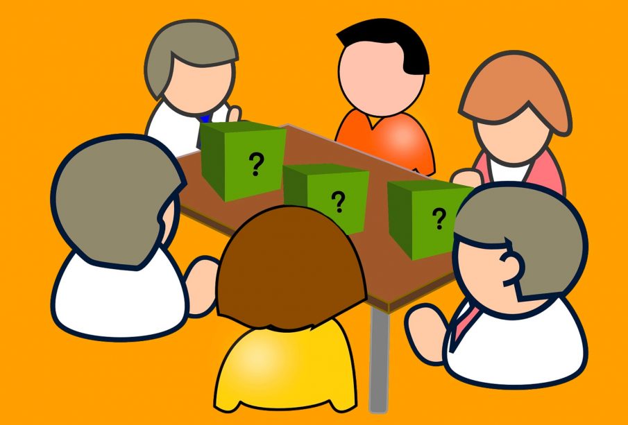 A group of people sit around a table with three boxes on the table surface and a question mark on each box.