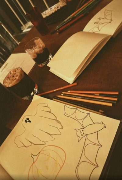 A photo of a table with two art filled notebooks (one near the camera and the other further down the table), coloured pencils strewn across the table, two dark beverages with ice in whiskey glasses in the middle of the table, and a deep red plastic cup at the far end of the table. The closer notebook has drawings of a ghost, a winking jack-o’-lantern, and a smiling bat while the further notebook has an unfinished drawing of a bat.
