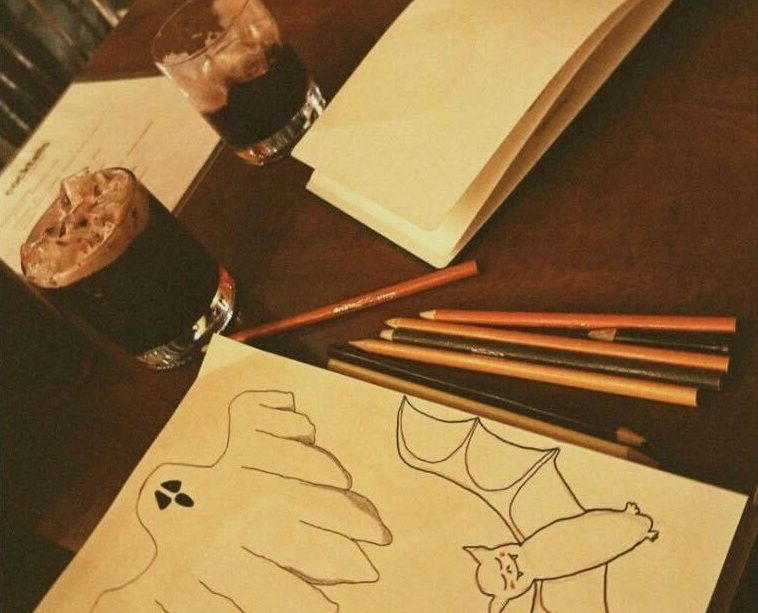 A photo of a table with two art filled notebooks (one near the camera and the other further down the table), coloured pencils strewn across the table, two dark beverages with ice in whiskey glasses in the middle of the table, and a deep red plastic cup at the far end of the table. The closer notebook has drawings of a ghost, a winking jack-o’-lantern, and a smiling bat while the further notebook has an unfinished drawing of a bat.