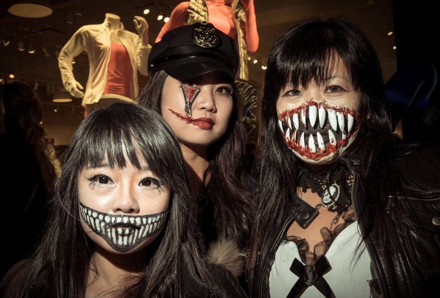 Three people with Halloween themed make-up in a photograph. The one on the left side has drawn a smiling mouth with sharp teeth onto her face, the one in the middle has a zipper opening her cheek under her eye and a scar pulling the side of her mouth open by using liquid latex, and the one on the right has a liquid latex and fake sharp tooth-ed mouth stretching across her face and covering her nose.