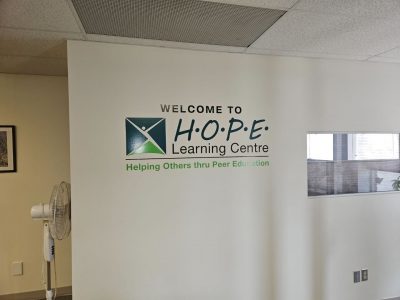 A white wall with the phrase “Welcome to HOPE Learning Centre” in black words with the phrase “Helping Others thru Peer Education” beneath it.