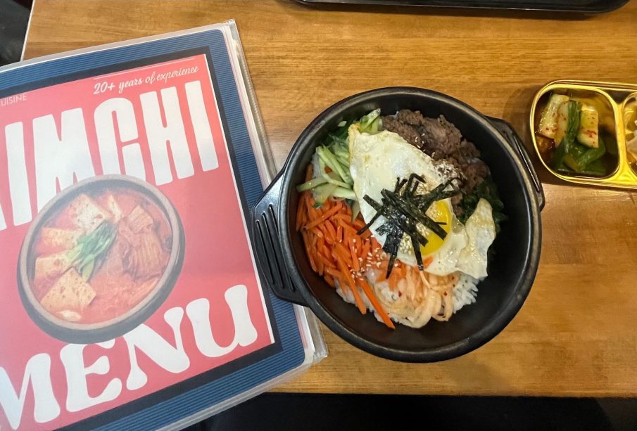 A photo of the front of a menu that reads “Kimchi Menu” and a meal. The meal consists of banchan in a stone bowl and a side of seasoned cucumbers and apple slices.