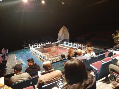 A photo of a stage set up with a fence around it, a gangway to the back right corner leading into a sheet doorway, and various trunks around the gangway and inside the fence.