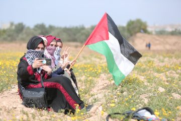 Four people sit in a field of dandelions wearing traditional Palestinian clothing. The third person of the four holds a Palestine flag out to billow in the wind.