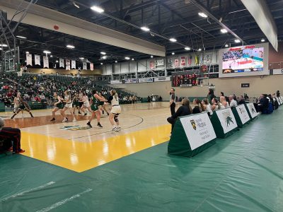 A photo of the University of Regina and Fraser Valley’s women’s basketball teams playing a match. The players are focused on the basketball in the air. The score is 33:25 in Fraser Valley’s favour.