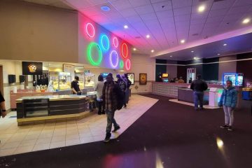 An image of the concession area of Moonlight Movies. There are some people wandering and one person working the register behind the counter. There are some chairs and tables and some arcade games at the back of the image.