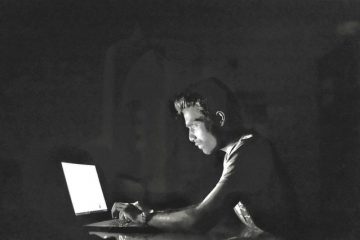 A person sitting in a dark room has their face illuminated by a laptop screen. They are typing intently.